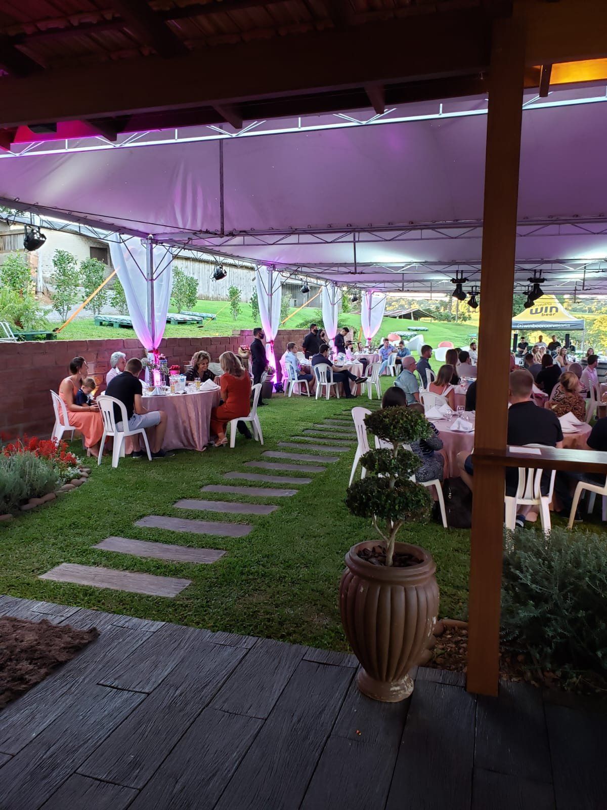 Festa à tarde sob tenda com iluminação rosa e mesas redondas