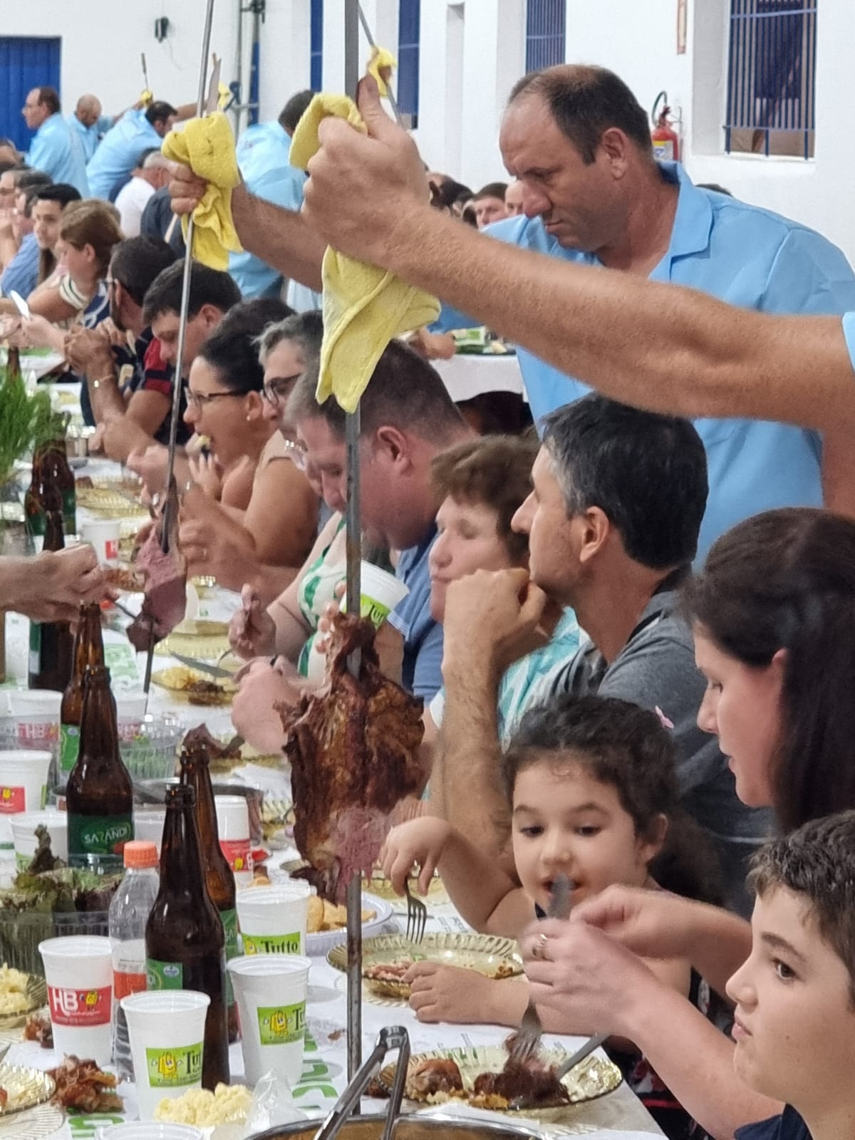 Churrasco sendo servido em mesa longa de festa comunitária